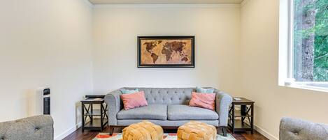 A living room with a grey sofa, two ottomans, a world map on the wall, and a window with a view of trees.