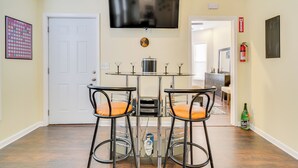 A dining area with a glass-top table, two bar stools with orange cushions, a flat-screen TV mounted above, and a fire extinguisher on the wall.