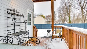 A covered outdoor deck with a metal bench and a wicker chair, a wooden railing, and a snow-covered ground.