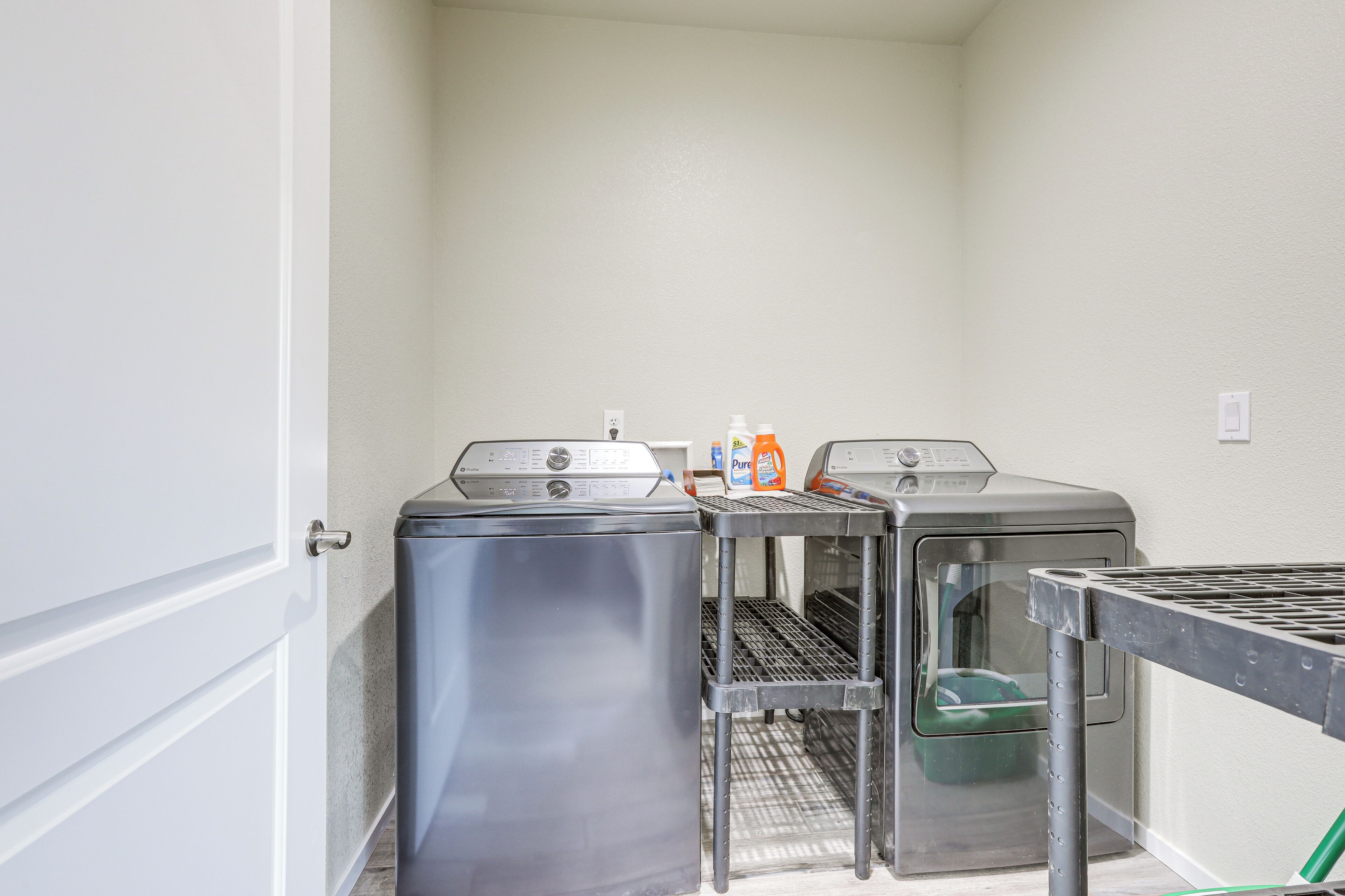 A laundry room with two stainless steel washers and dryers, a metal shelf, and a green trash bin.