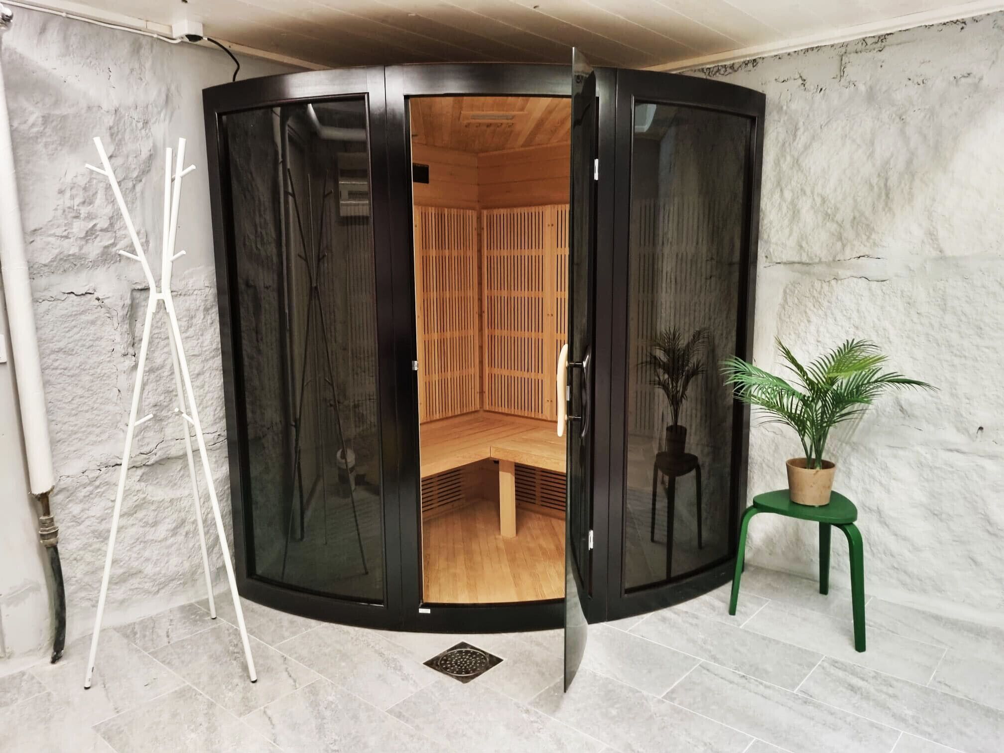 Une pièce dotée d’une porte en verre noir qui donne sur un espace sauna en bois. Un tabouret vert avec un pot de plante est placé à côté de la porte.