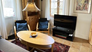 Una habitación con una mesa de comedor de madera, dos sillas, un televisor, un armario y un ventanal con cortinas.