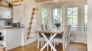 A dining area with a wooden table, white chairs, and a view of greenery through large windows.