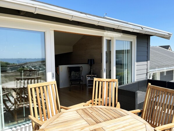 A wooden deck with chairs, a table, and a glass door leading to an interior space with a lamp and a chair.