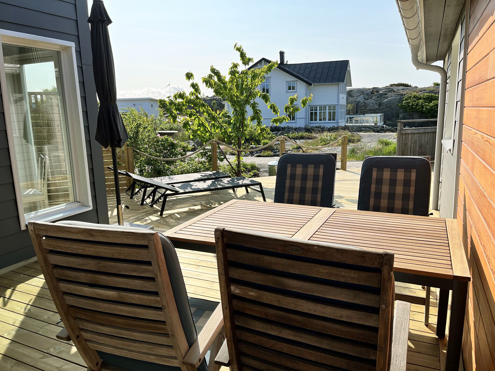 Une terrasse en bois avec une table et des chaises, une barrière en corde et une vue sur une maison et des arbres.