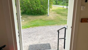 Eine Tür, die zu einem Kieshof mit einem Metallgitter führt, umgeben von Grünpflanzen und wolkenlosem Himmel.