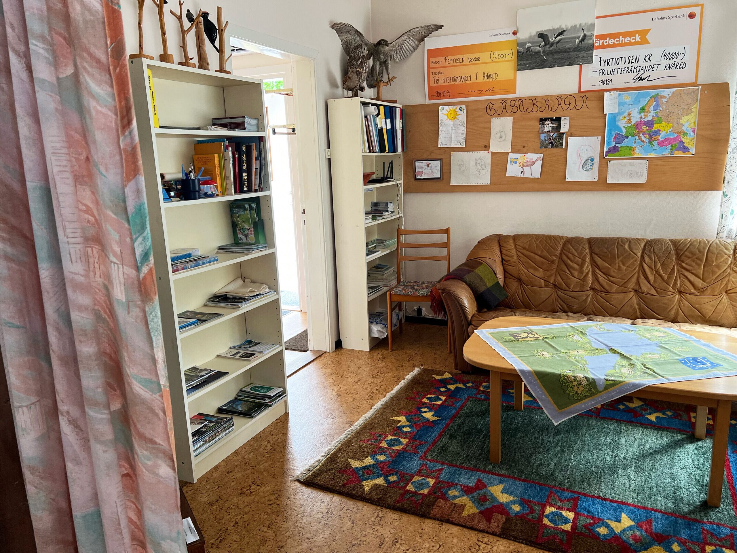 A cozy room with a brown leather sofa, a wooden chair, a small table with a map, and a bulletin board with papers and photos.