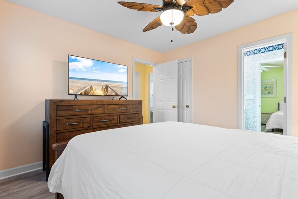 A bedroom with a bed, a wooden dresser, a ceiling fan, and a TV on the wall.