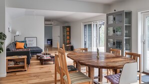 A spacious living area with a wooden dining table, comfortable seating, a bookshelf, and a view through a glass door.