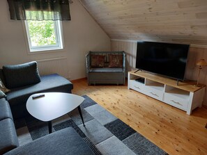 Ein gemütliches Wohnzimmer mit einer Couch, einem kleinen Tisch, einem Fernseher auf einem Holzschrank und einem Fenster mit Vorhängen.