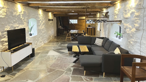 A modern living room with a stone floor, a black sofa, a wooden coffee table, a TV on a white cabinet, and a staircase leading to another room.