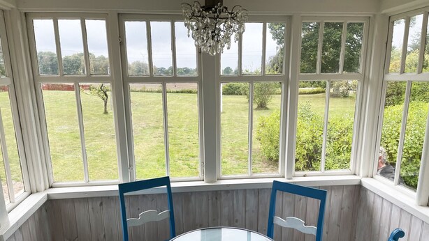 Un invernadero con una mesa redonda de cristal y sillas azules, ventanas amplias con vistas a un campo cubierto de hierba.