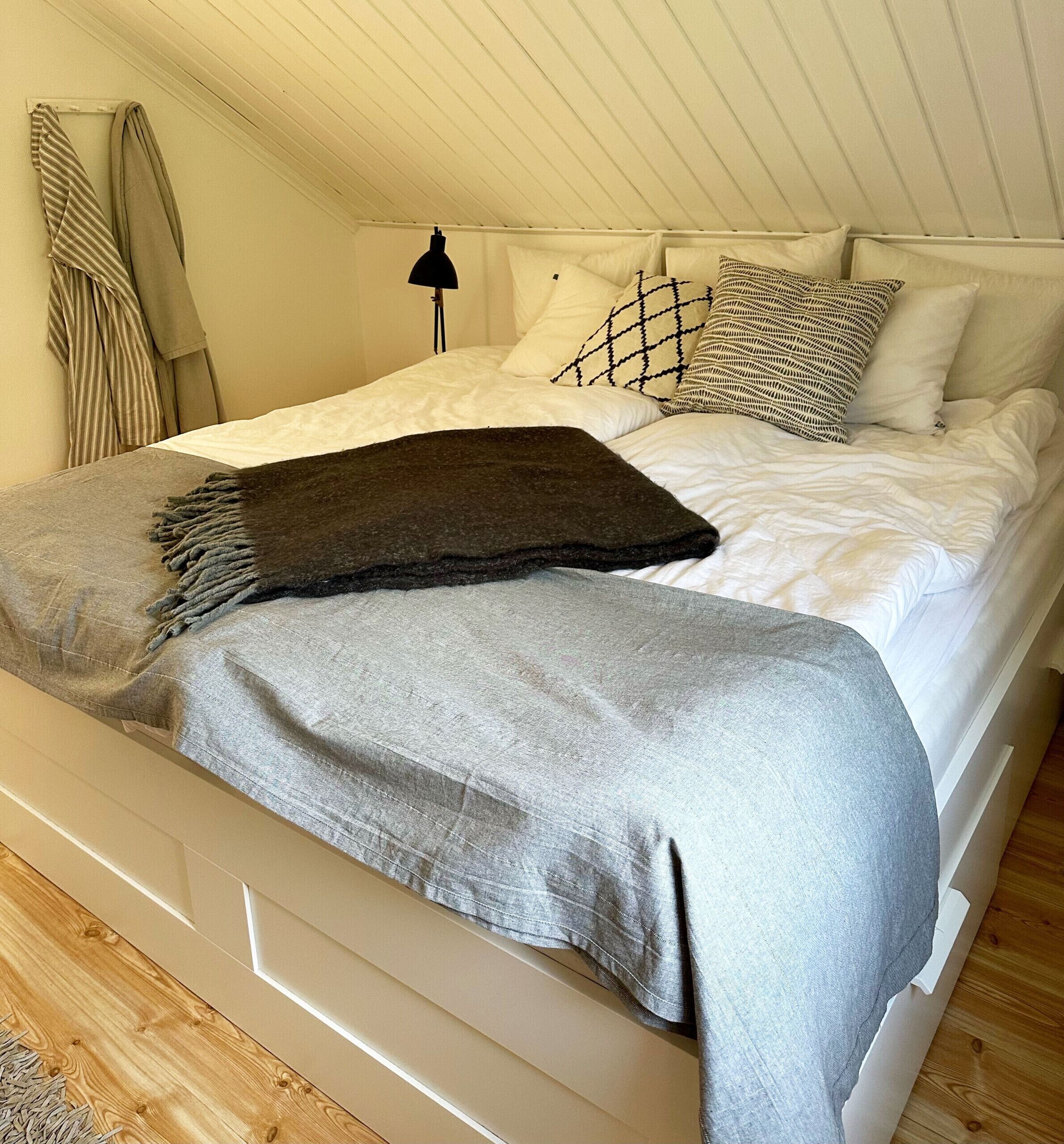A bed with a grey blanket, white bedding, and patterned pillows in a room with wooden flooring and a sloped ceiling.