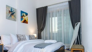 Quarto de hotel com uma cama, duas pinturas na parede, uma janela grande com cortinas e uma mesinha.