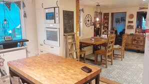Un comedor con mesa y sillas de madera, una cocina con horno, una pizarra con escritura y una araña.