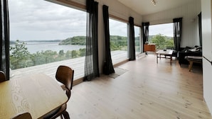 A spacious room with large windows, a wooden floor, a dining table, a sofa, and a small table with a lamp.