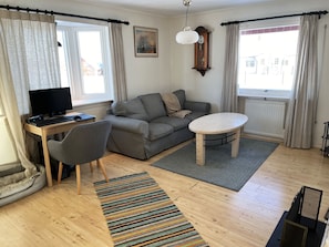 Een woonkamer met een bank, een bureau met een computer, een stoel, een ronde tafel, een klok en een raam met gordijnen.
