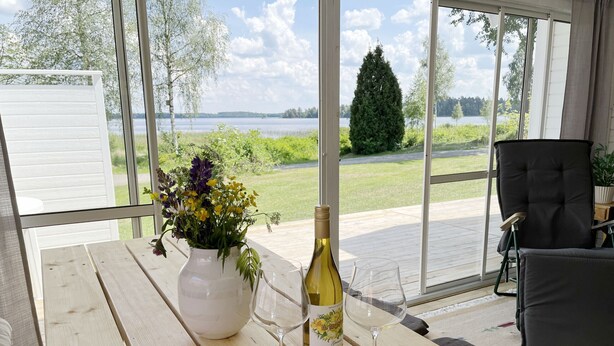 A wooden table with a bottle of wine and two glasses, a vase of flowers, and a view of a lake and greenery through large windows.