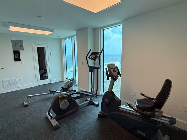 A gym with cardio machines, a standing bike, and a treadmill, with a large window overlooking the ocean.