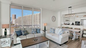 A modern living room with two white sofas, a coffee table, and a dining area with a white table and chairs. There is a large window with a view of high-rise buildings. - Perdido Dunes Tower 7W by Liquid Life (Orange Beach)