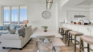 A modern kitchen with white cabinets, a large island with stools, and a dining area with a white table and chairs. - Perdido Dunes Tower 7W by Liquid Life (Orange Beach)