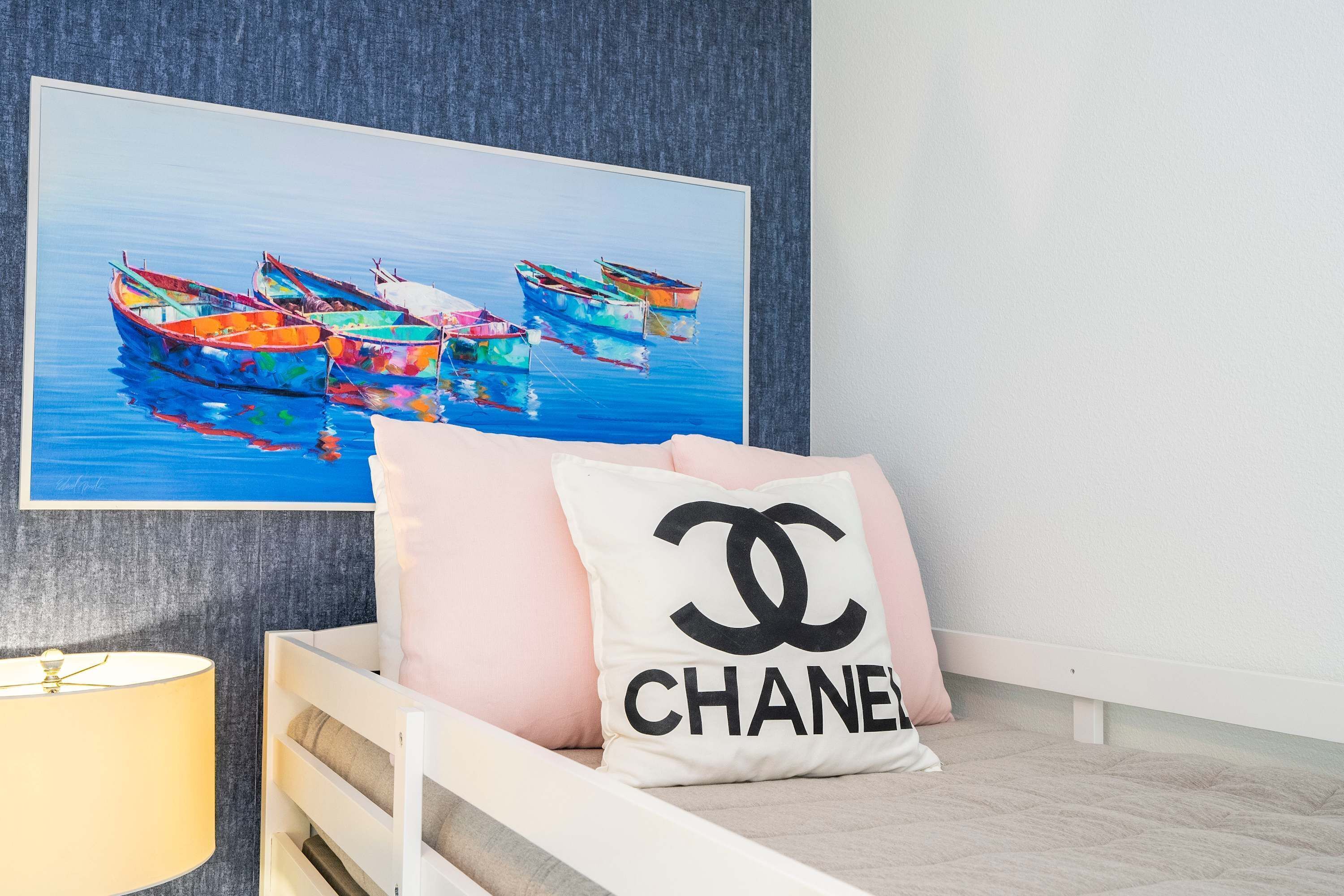 Une chambre à coucher avec un lit, un oreiller orné du logo Chanel, un tableau représentant des bateaux et une lampe.