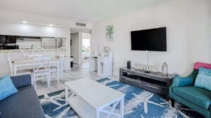 Ein modernes Wohnzimmer mit Sofa, Couchtisch und Fernseher. Es gibt eine Essecke mit Stühlen und Tisch. Der Raum hat weiße Wände und einen blauen Teppich.
