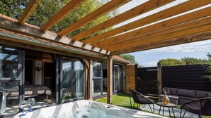 A hot tub under a wooden pergola with glass doors leading to a bedroom. - Romantic Countryside Lodge With Hot Tub (Chichester)