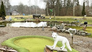 Un jardín ajardinado con una estatua de un gato blanco sobre una roca, un estanque y una casa al fondo.