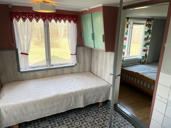 Une petite pièce avec un lit simple, une fenêtre avec des rideaux, une porte donnant sur une autre pièce et un sol à motifs.