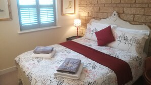 A bedroom with a brick wall, a window with blinds, a bed with floral bedding, a red throw blanket, and a bedside lamp.