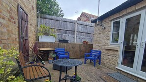 A patio area with a brick floor, a round table, two blue chairs, a wooden bench, and potted plants.