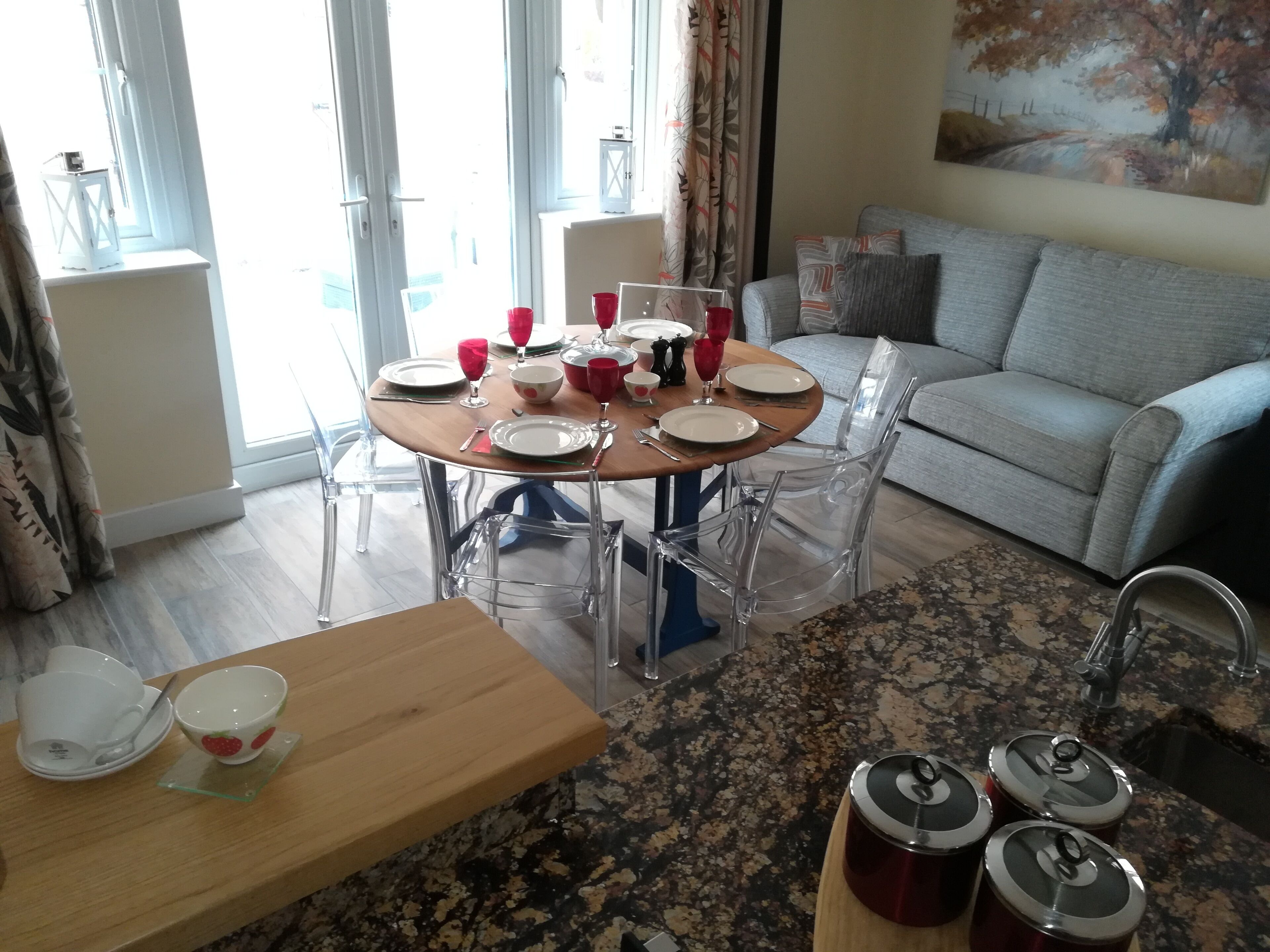A dining area with a round table set for four, clear chairs, a wooden table, and a kitchen area with granite countertops and stainless steel appliances.