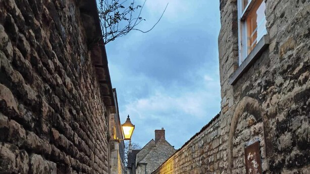 A narrow stone alleyway with brick flooring, flanked by stone walls and a single lit street lamp.