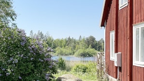 Un edificio rojo con base de piedra, un pequeño jardín con flores moradas y un lago al fondo.