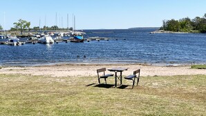 Un puerto deportivo con barcos amarrados, un área verde con una mesa y sillas, y una vista despejada del agua y la costa lejana.