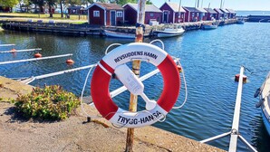 A lifebuoy with text "REVSUDDENS HAMN" and "TRYG-HANS" on a dock by a calm water body with boats and red buildings in the background.