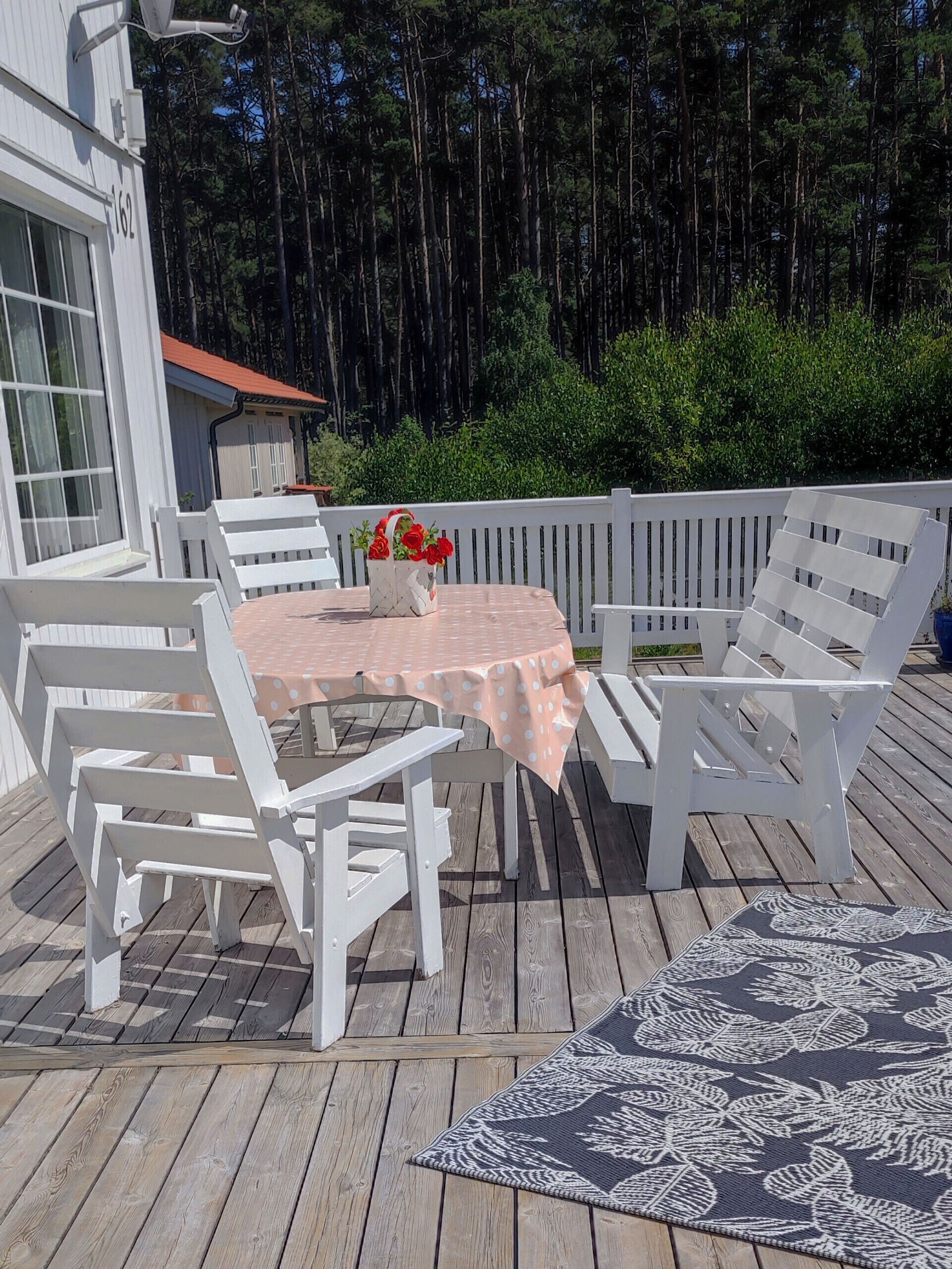 Une terrasse en bois, avec des chaises blanches et une table, entourée d’une clôture blanche et de verdure.