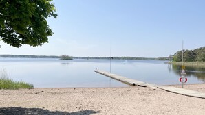A sandy beach with a wooden dock extending into calm water, surrounded by trees and a clear sky.