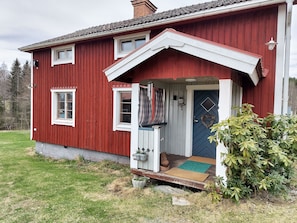 Ett rött trähus med en blå dörr, en liten veranda och en grön buske framför.