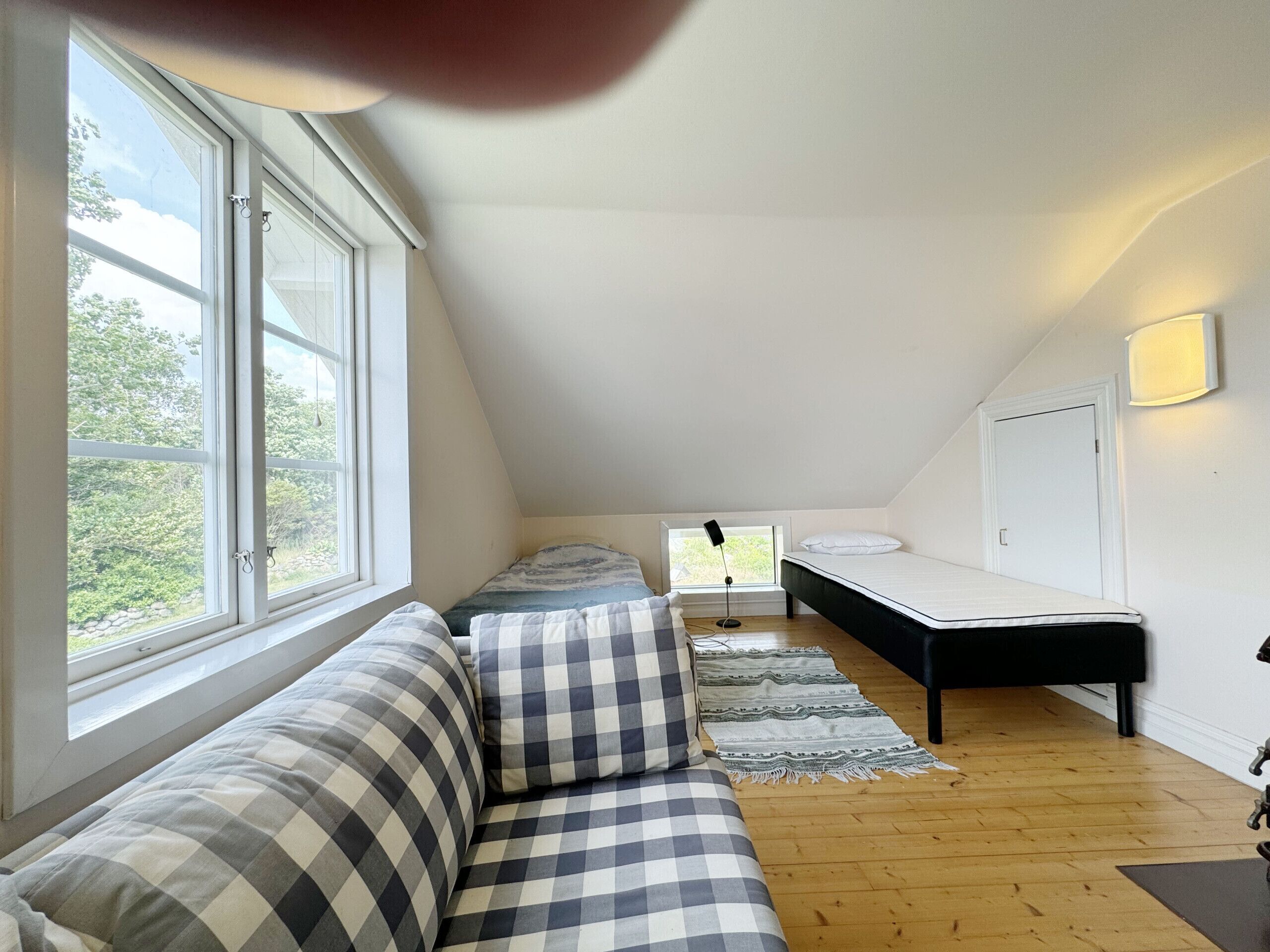 A cozy attic bedroom with a checkered sofa, a bed, a small desk, and a window with a view of greenery.