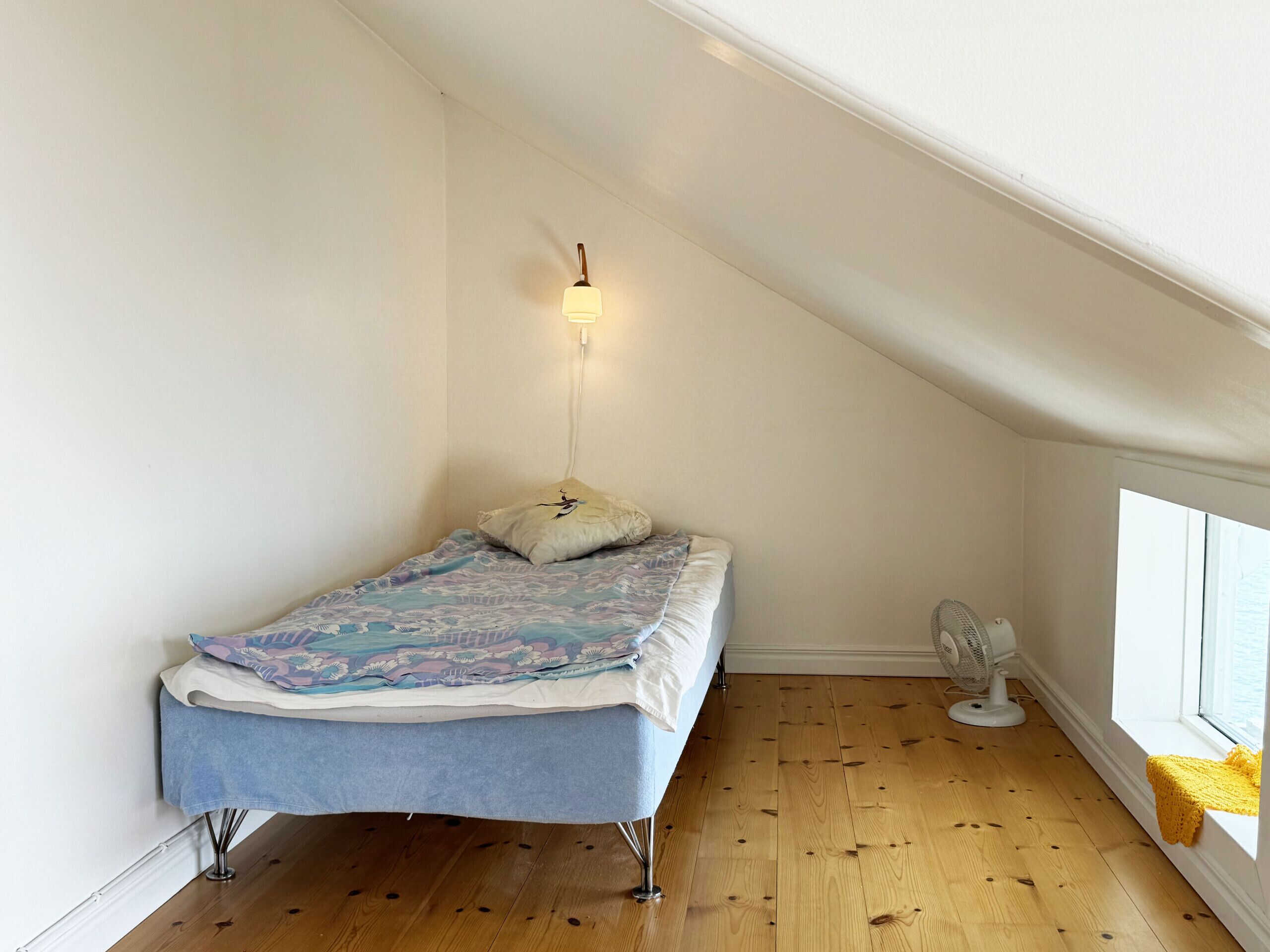 Quarto pequeno com uma cama de solteiro, teto inclinado, um ventilador e uma janela com um cobertor amarelo.