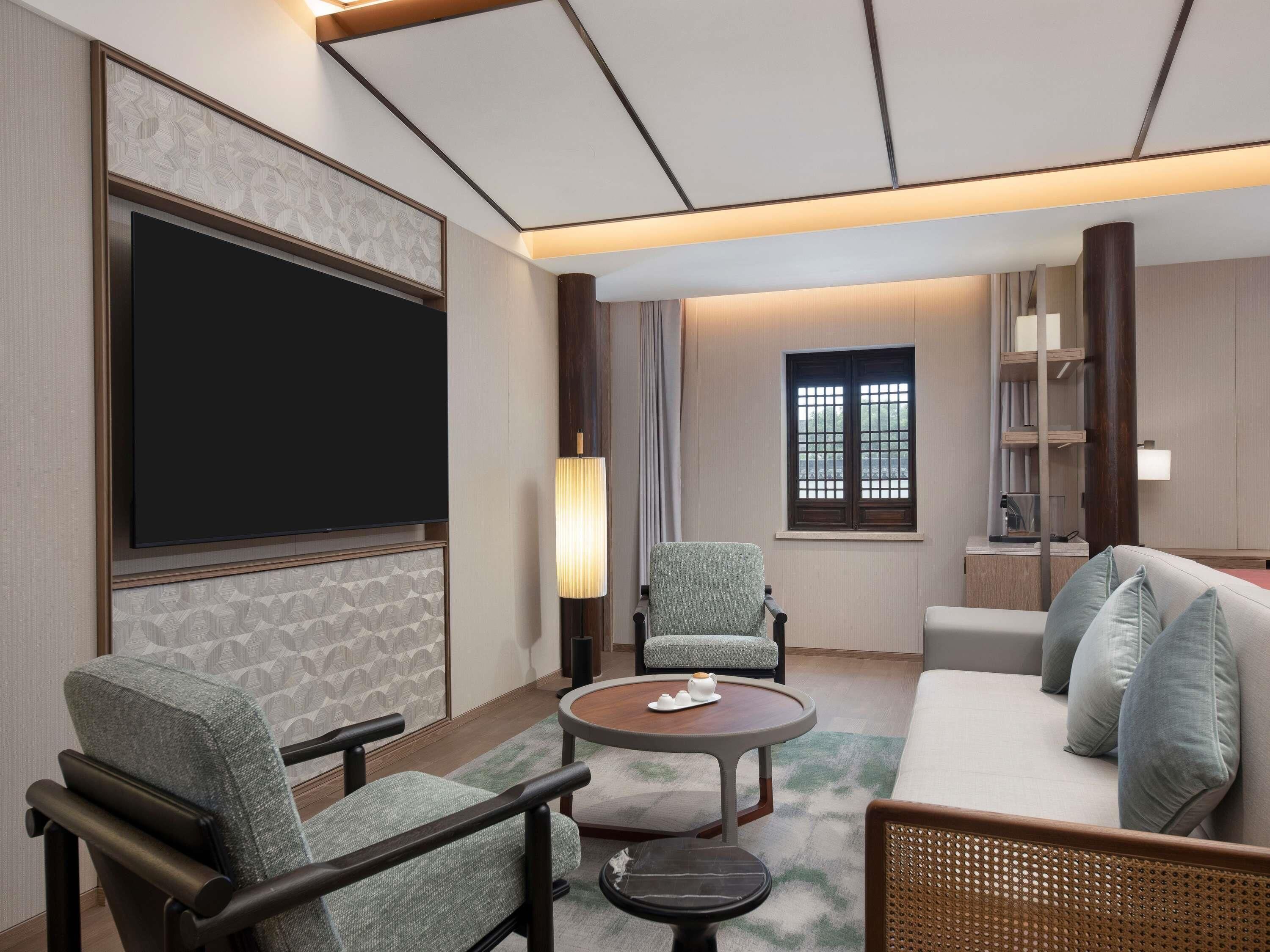 A hotel room with a large flat-screen TV, a sofa, two armchairs, a small round table, and a window with a view.
