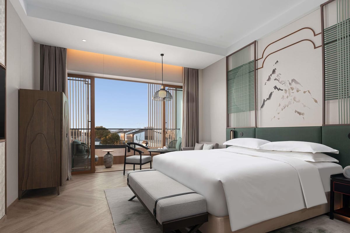 Een moderne hotelkamer met een groot bed, een zithoek met een tafel en uitzicht op de stad door grote ramen.