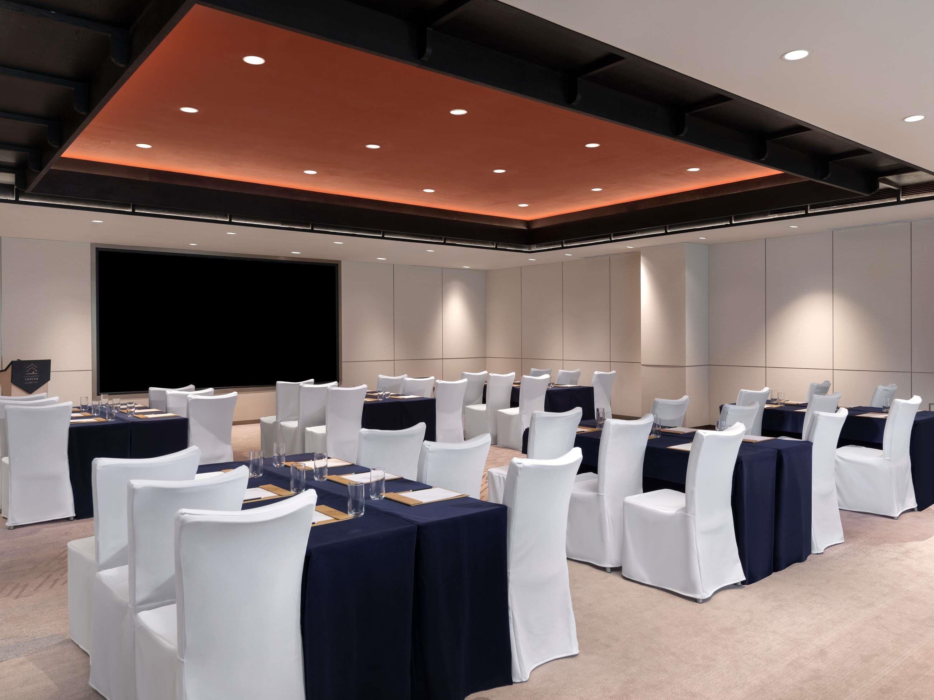 A conference room with rows of white chairs, dark blue tables, and a large screen.