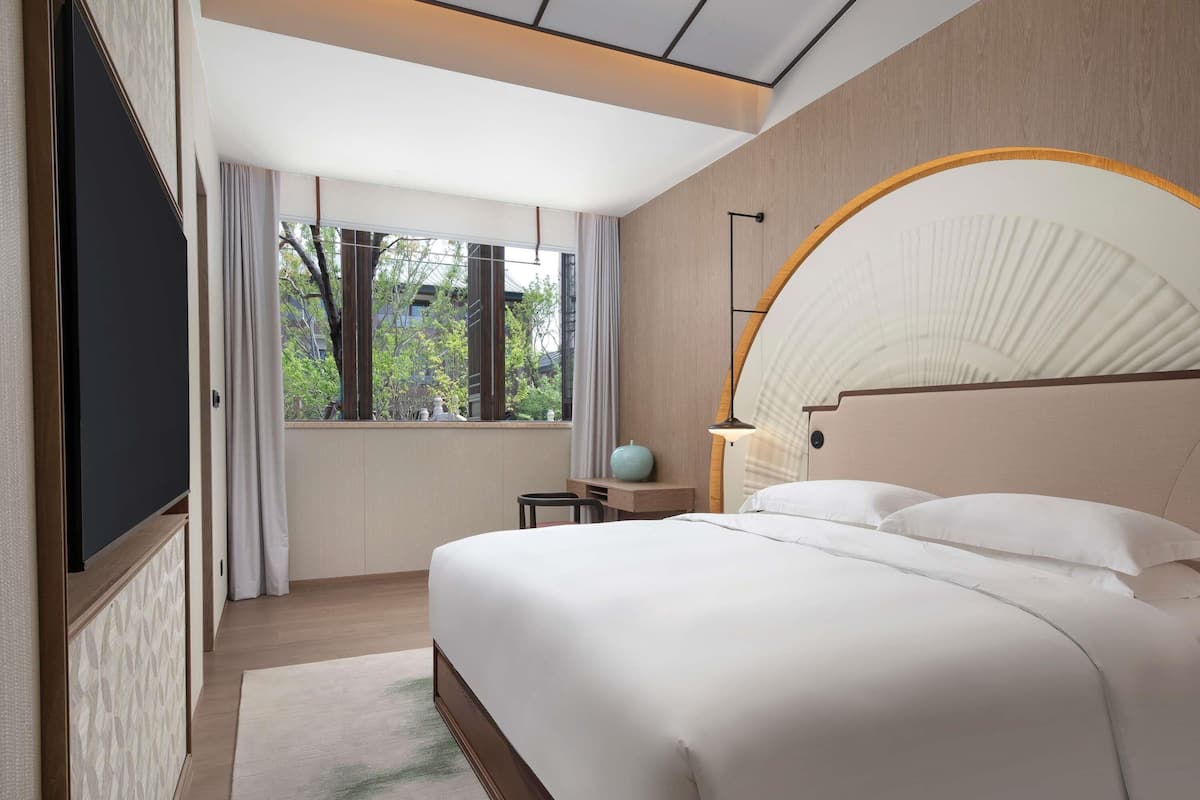 Een moderne hotelkamer met een groot bed, een televisie, een raam met uitzicht op groen en een opvallend gebogen hoofdeinde.