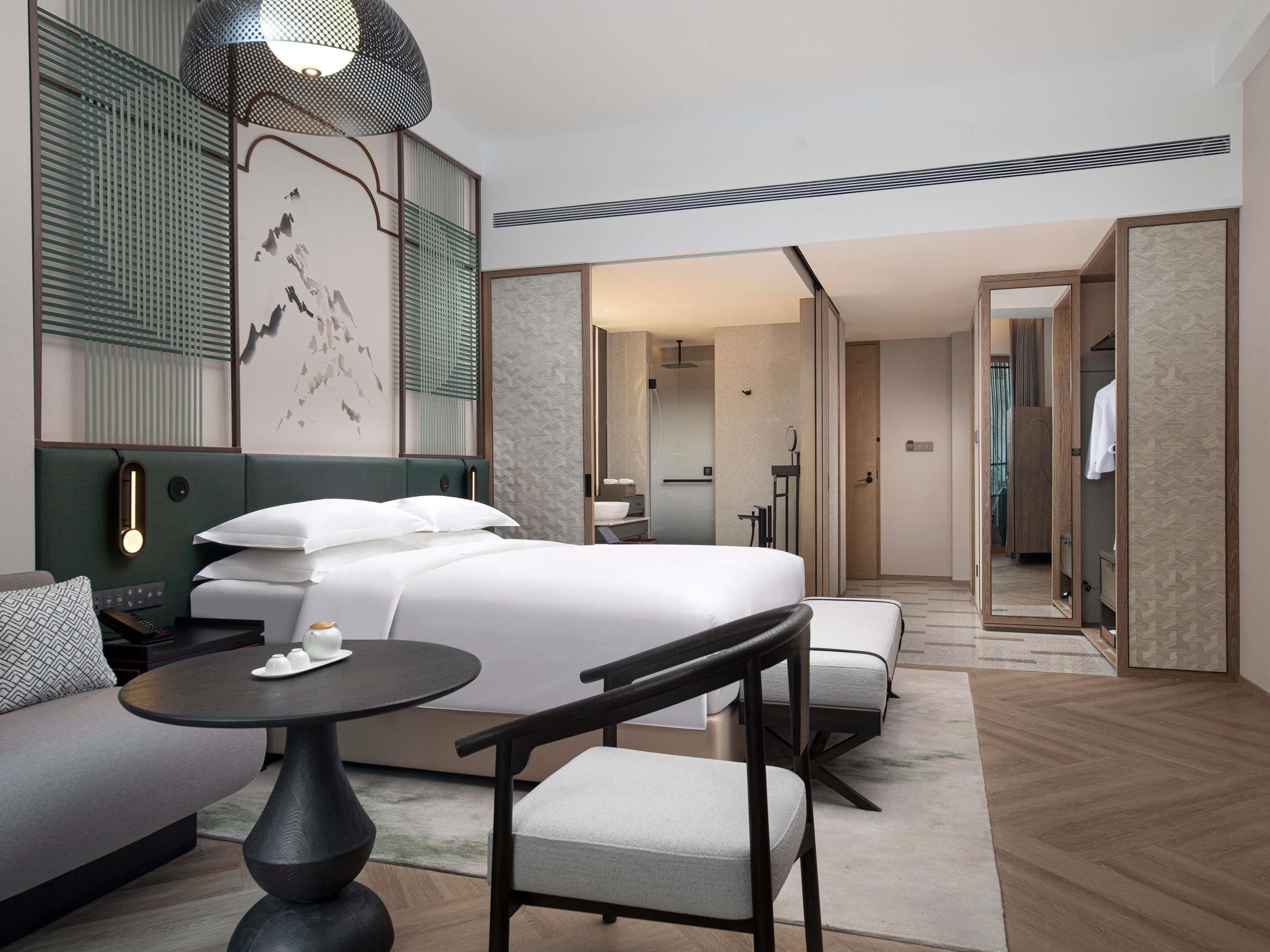 Una habitación de hotel moderna con una cama grande, una zona de estar con una mesa y sillas, un espejo y un vestidor.