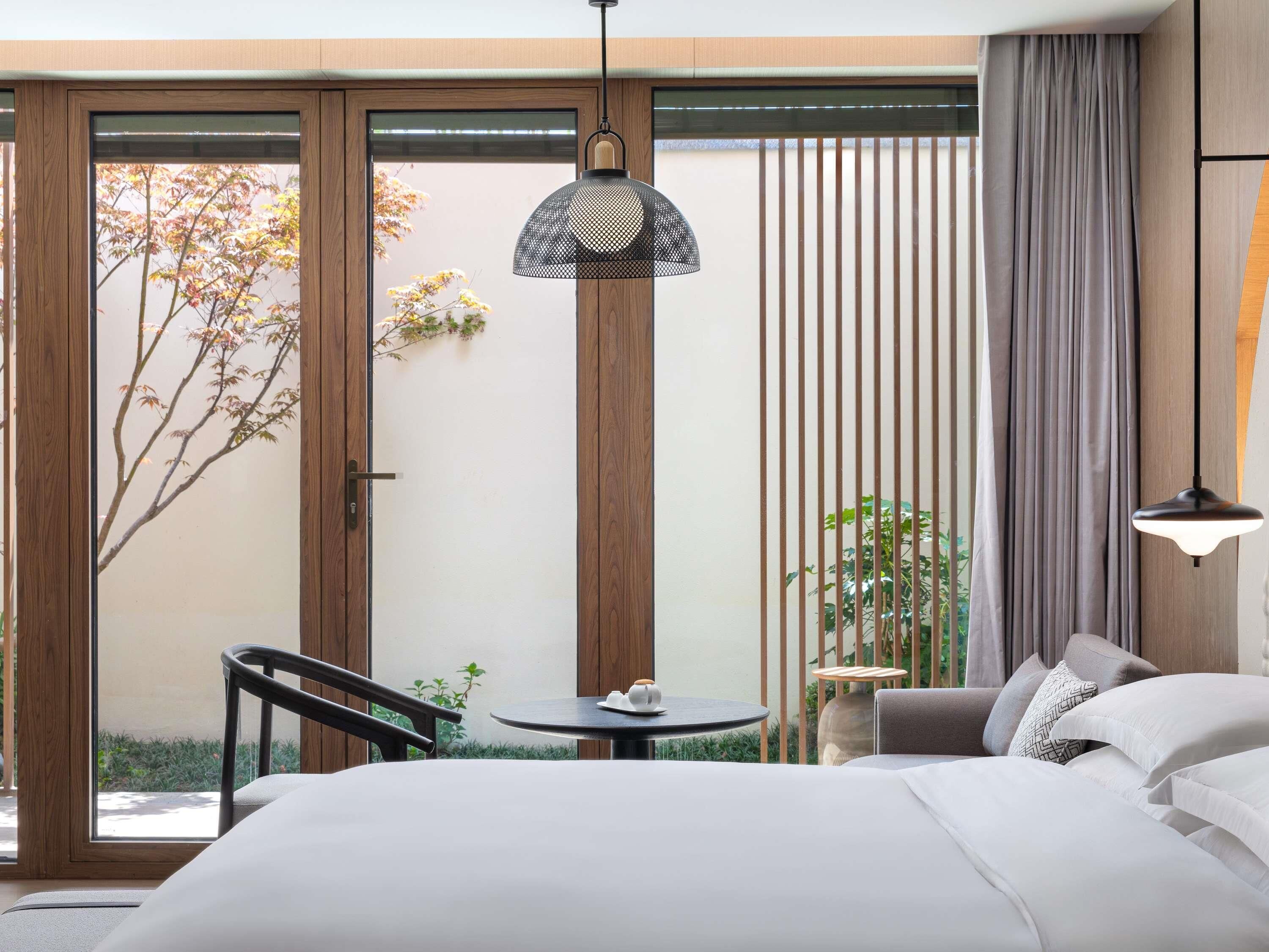 A hotel room with a bed, a round table, a chair, a hanging lamp, and a view of a garden through a sliding door.