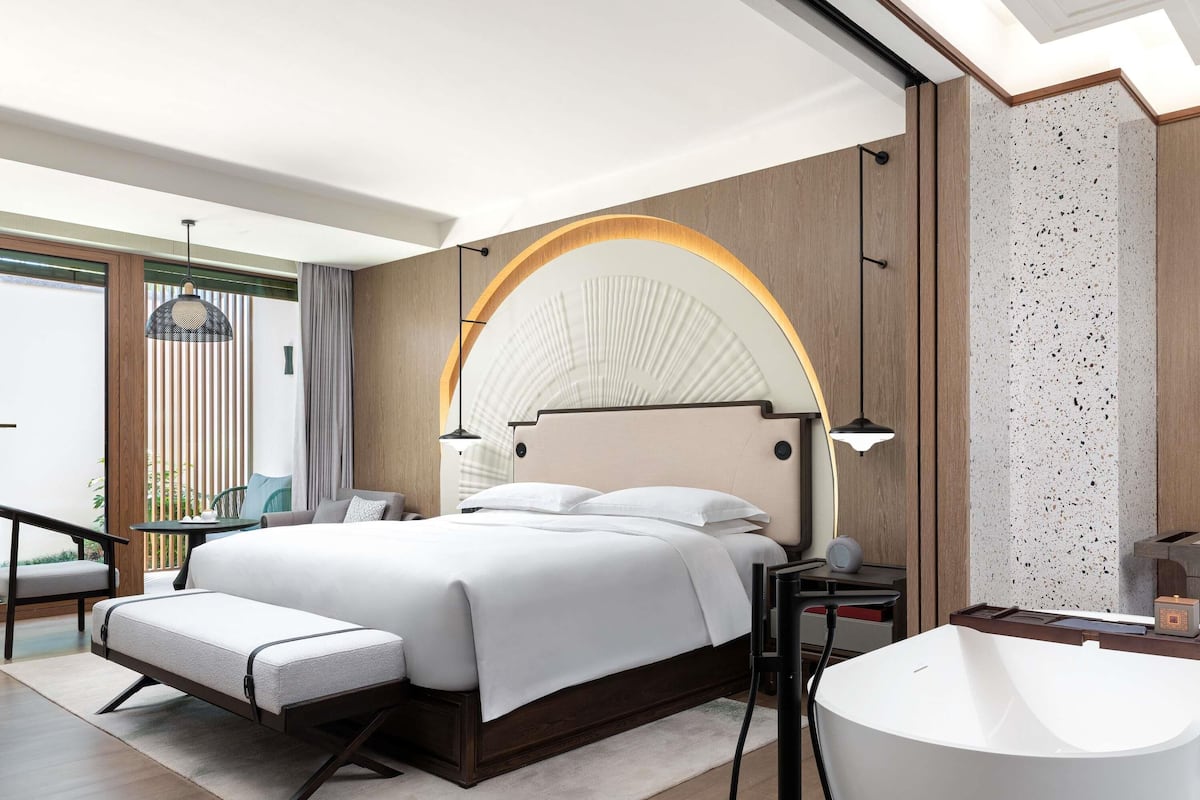 Een moderne hotelkamer met een groot bed, een zithoek met een stoel en een tafel, een badkuip en uitzicht op groen.