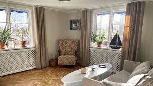 Una sala de estar acogedora con un sillón estampado, una mesita de centro blanca y un sofá. Hay plantas en macetas en el alféizar de la ventana y un pequeño modelo de un velero en la ventana.